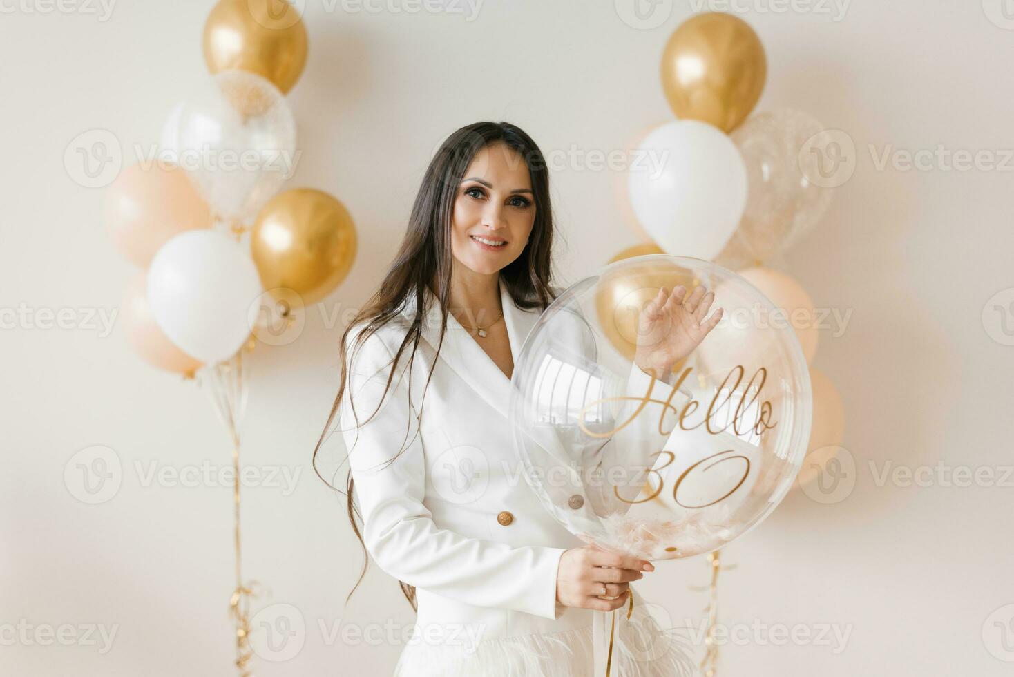 positief vrouw vieren verjaardag gekleed in elegant veer jurk poseren met verjaardag ballon in de omgeving van opgeblazen ballonnen Bij huis foto