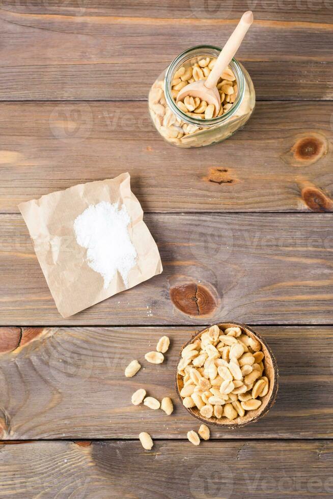 gezouten pinda's in een kom en in een pot met een lepel en zout Aan papier Aan een houten tafel. gezond vegetarisch voedsel. rustiek stijl. verticaal en top visie foto