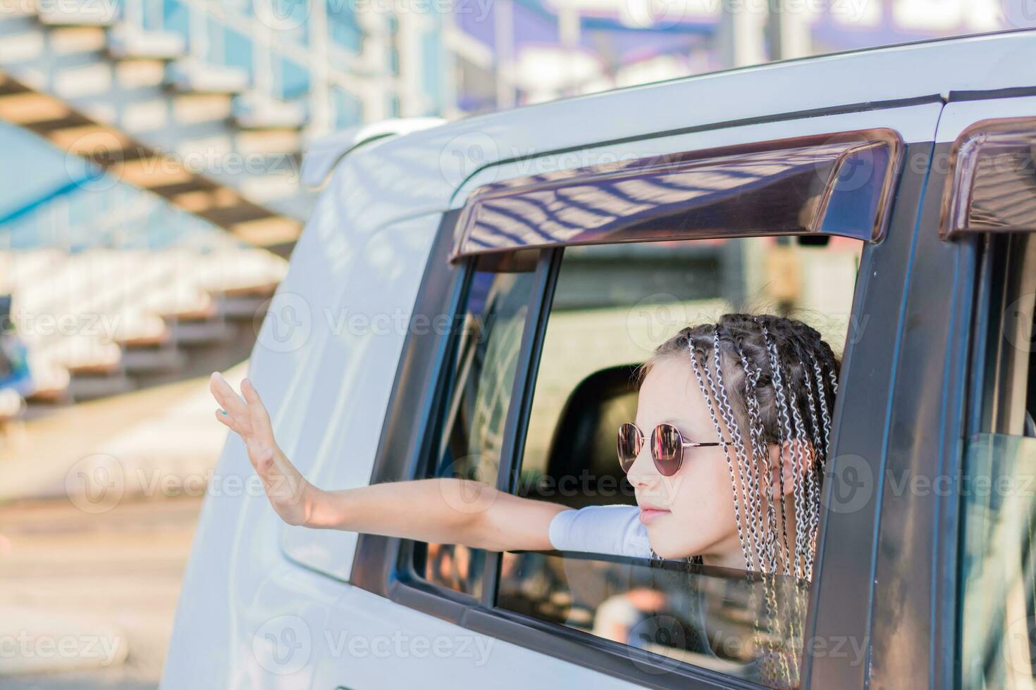 een schattig tiener- meisje in zonnebril met afro-vlechten golven haar hand- uit de venster terwijl zittend in de auto. weg reis foto