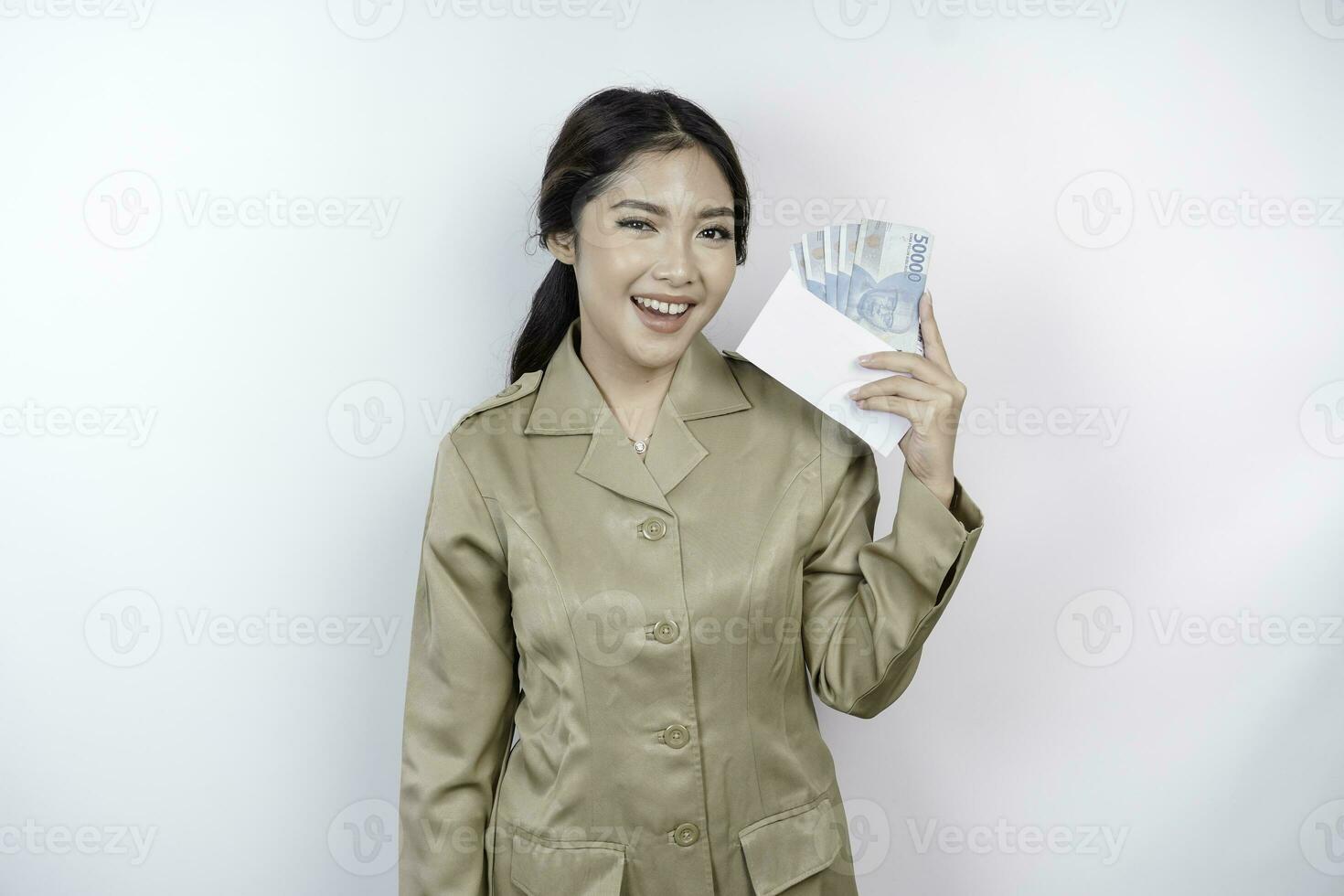 een glimlachen jong regering arbeider vrouw is vervelend khaki uniform Holding contant geld geld in Indonesisch roepia geïsoleerd door wit achtergrond foto