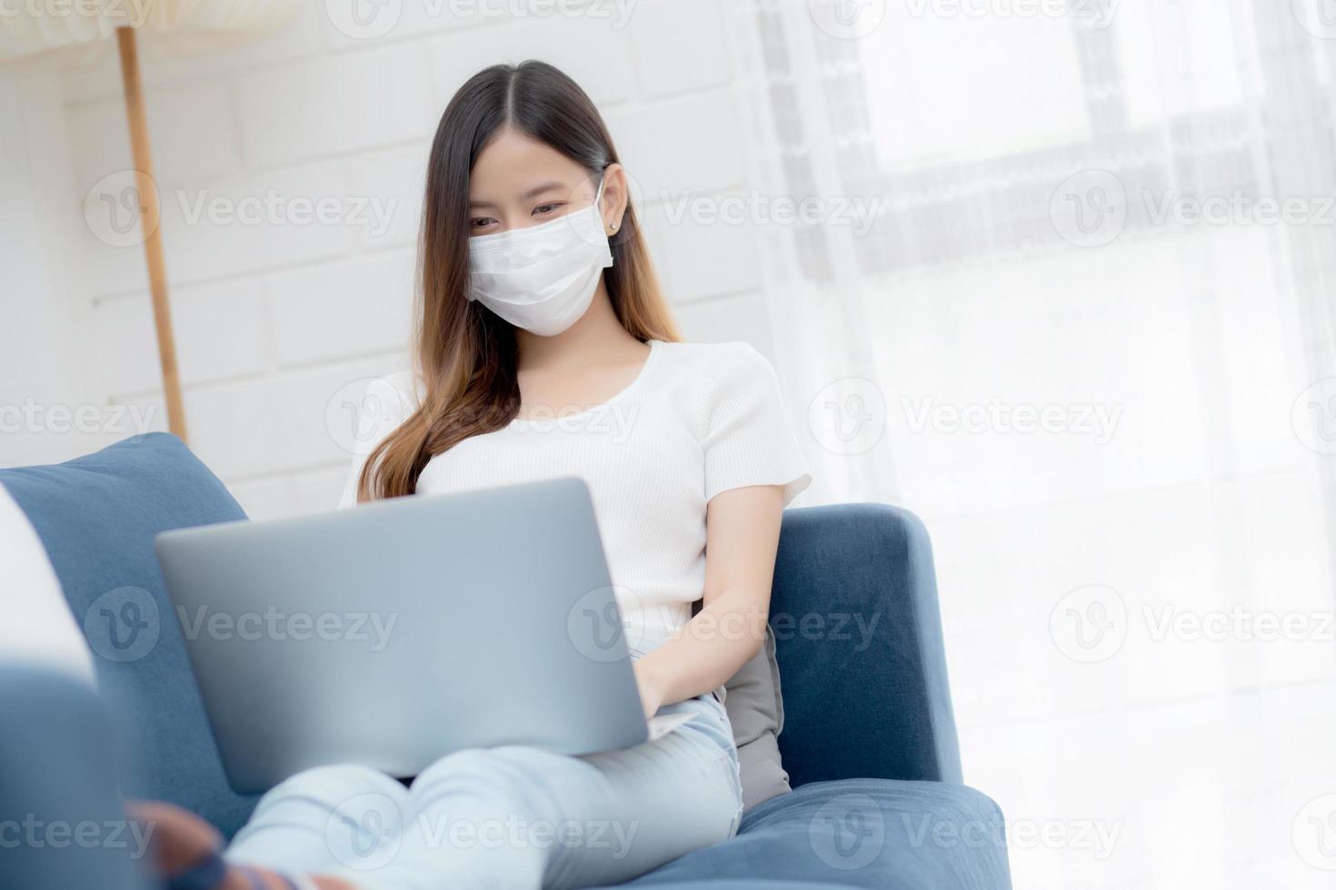 jonge aziatische zakenvrouw met gezichtsmasker werkt vanuit huis op laptopcomputer online naar internet in de woonkamer, meisje met medisch masker en quarantaine voor pandemie van covid-19, blijf thuis, nieuw normaal. foto