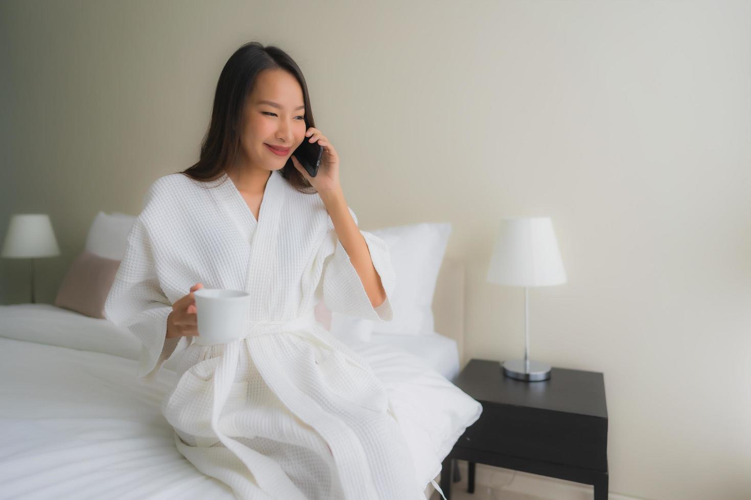 portret mooie jonge Aziatische vrouwen met koffiekopje en mobiele telefoon op bed on foto