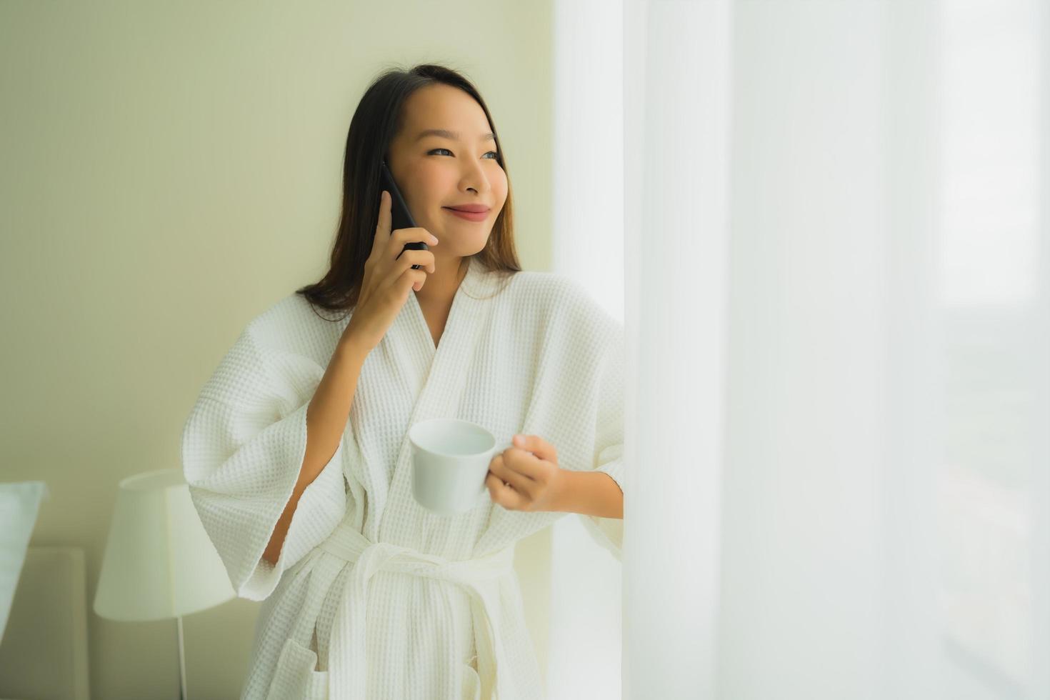 portret mooie jonge Aziatische vrouwen met koffiekopje en mobiele telefoon op bed on foto