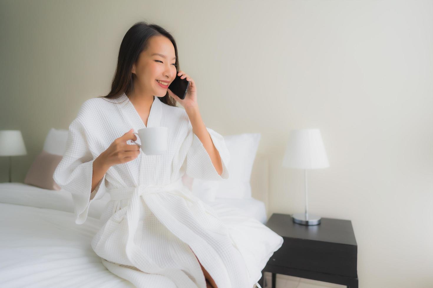 portret mooie jonge Aziatische vrouwen met koffiekopje en mobiele telefoon op bed on foto