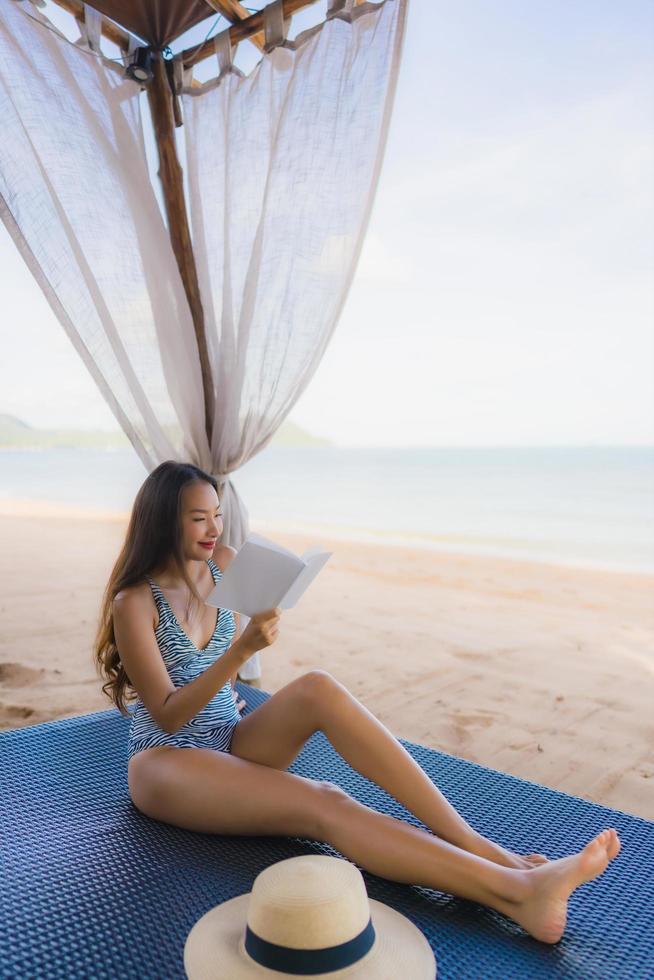 portret mooie jonge aziatische vrouw die een boek leest met een gelukkige glimlach ontspan in een lounge bed stoel op het strand zee oceaan voor vrije tijd foto