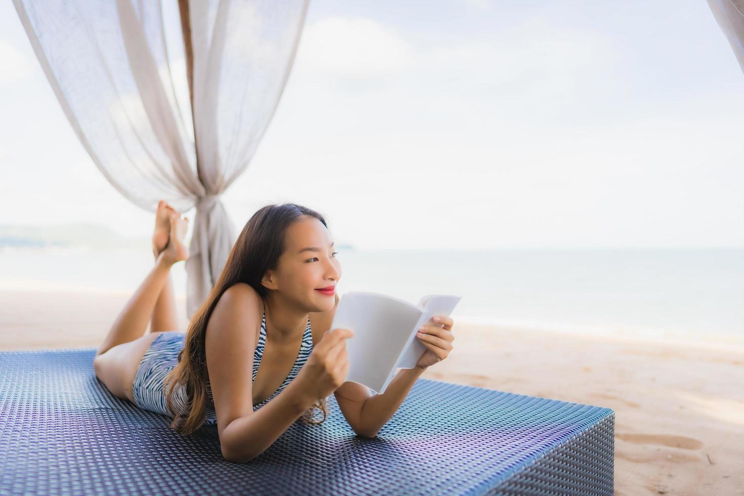 portret mooie jonge aziatische vrouw die een boek leest met een gelukkige glimlach ontspan in een lounge bed stoel op het strand zee oceaan voor vrije tijd foto