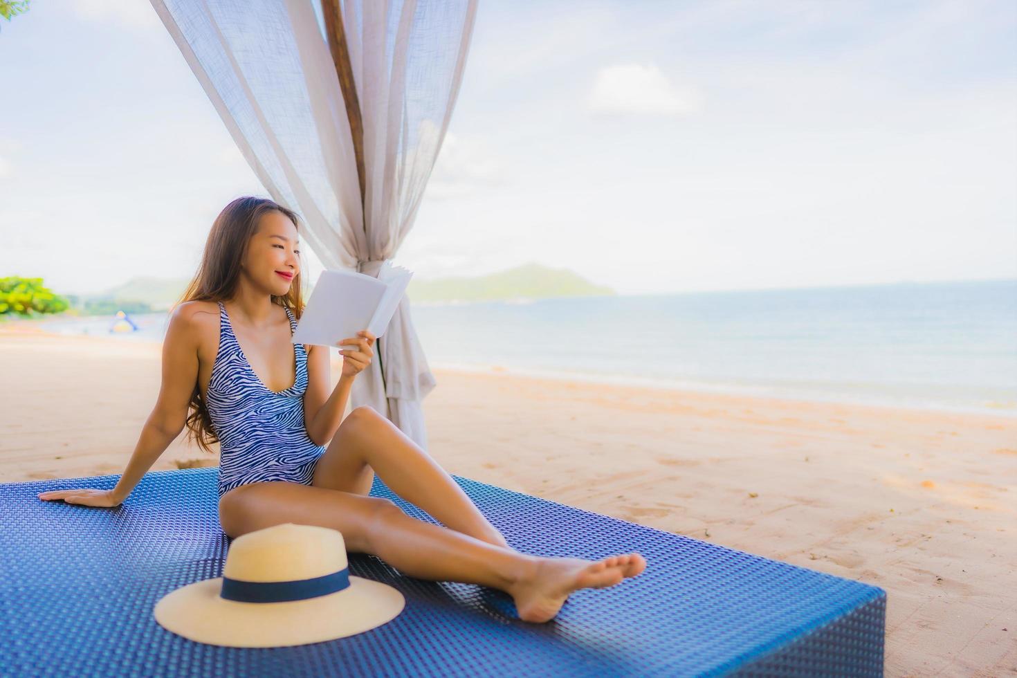 portret mooie jonge aziatische vrouw die een boek leest met een gelukkige glimlach ontspan in een lounge bed stoel op het strand zee oceaan voor vrije tijd foto