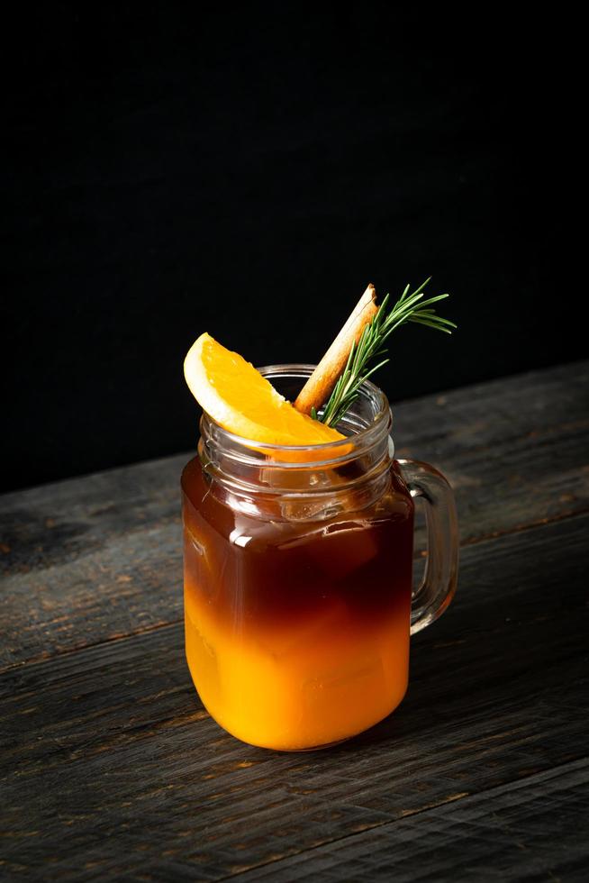 een glas iced americano zwarte koffie en een laagje sinaasappel- en citroensap versierd met rozemarijn en kaneel op een houten ondergrond foto