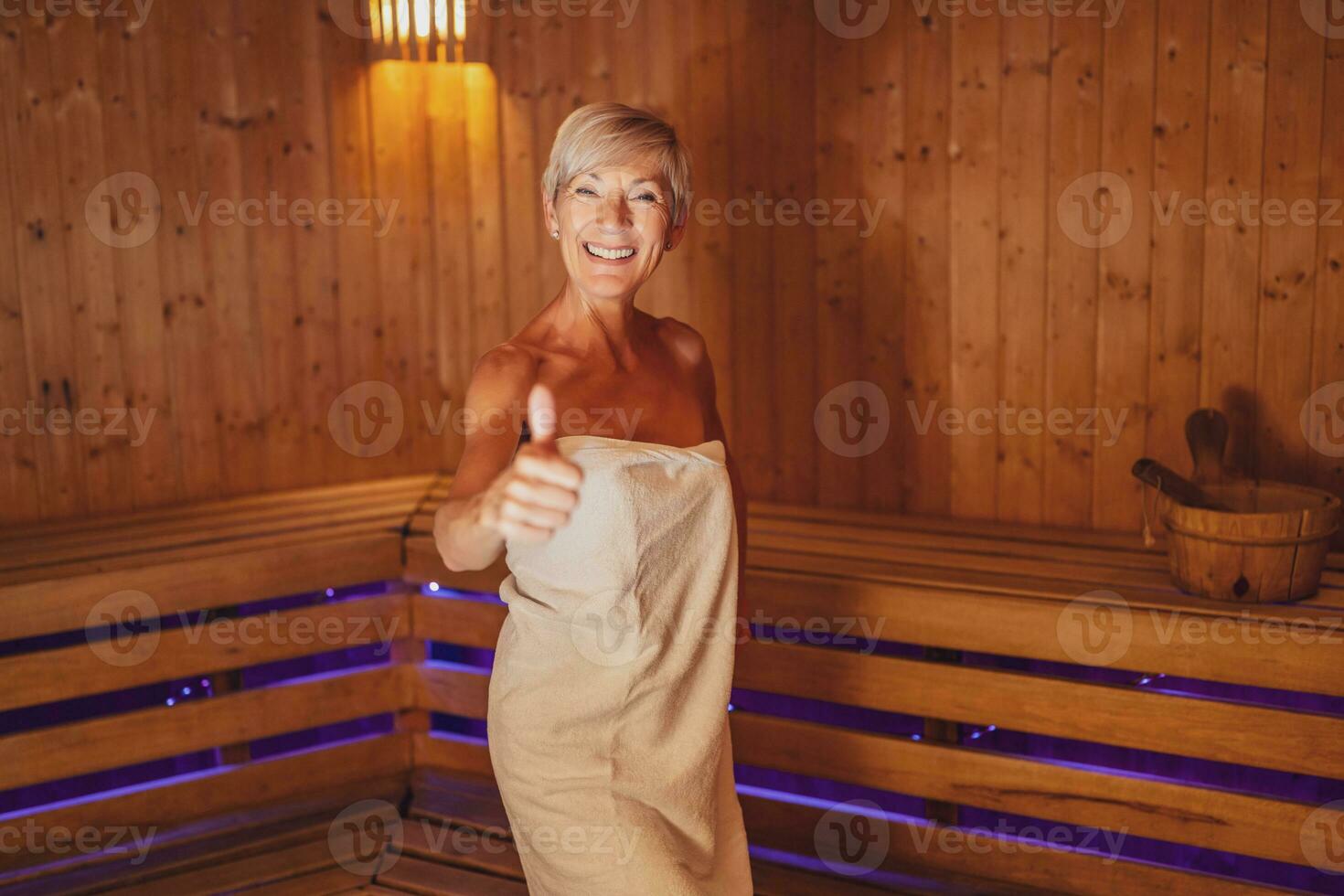 volwassen vrouw is klaar voor ontspannende in sauna. ze is staand in sauna en tonen duimen omhoog. spa concept. foto