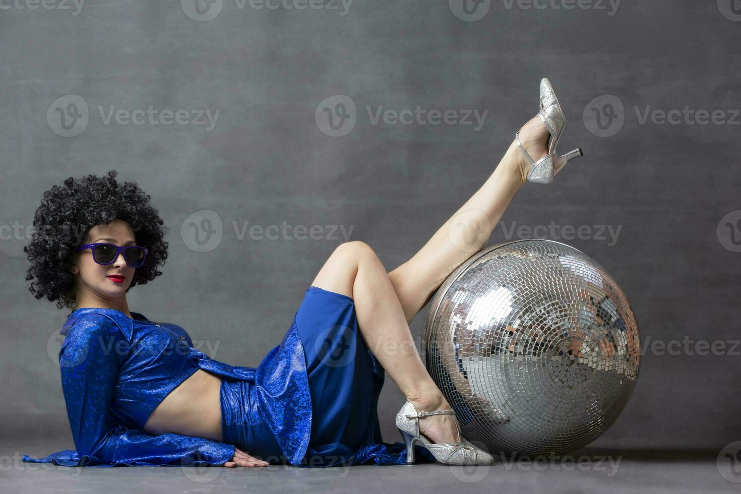 meisje met een disco bal in een afro pruik Aan een grijs achtergrond. disco stijl van de jaren zeventig of jaren tachtig. foto