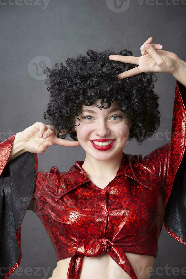 een mooi meisje in een rood disco kostuum en een zwart gekruld pruik glimlacht. foto