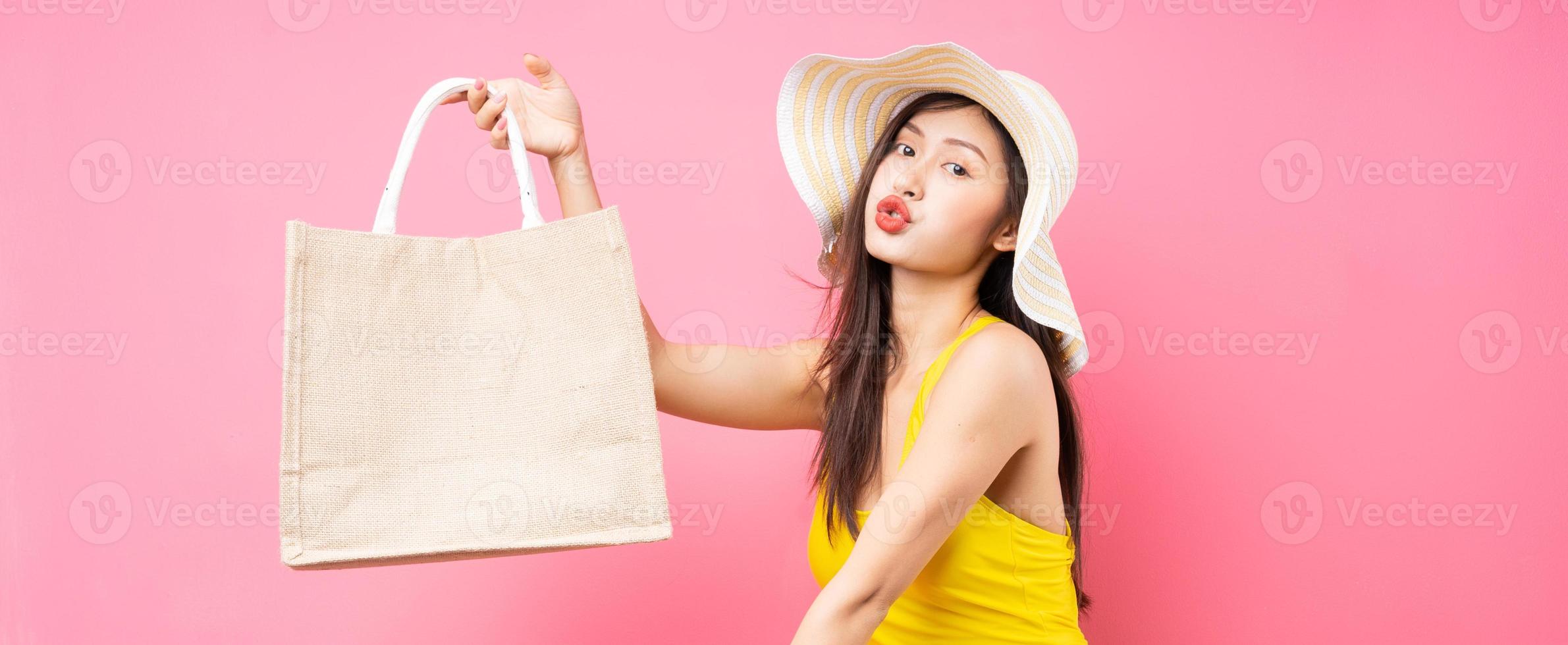 mooie jonge aziatische vrouw in geel zwempak, tas en breedgerande hoed die zich voordeed op roze achtergrond foto