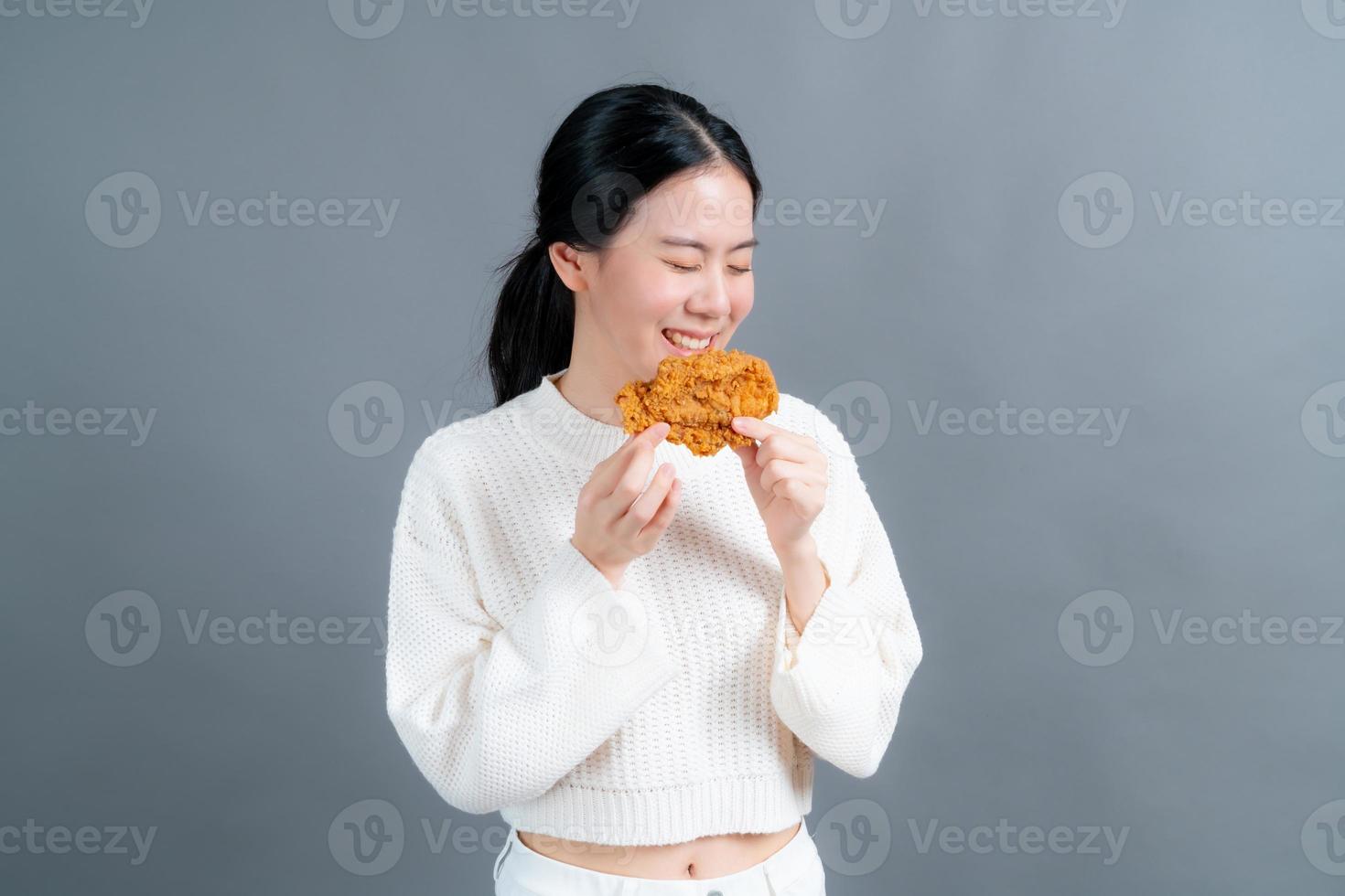 jonge aziatische vrouw die een trui draagt met een blij gezicht en geniet van het eten van gebakken kip op een grijze achtergrond foto