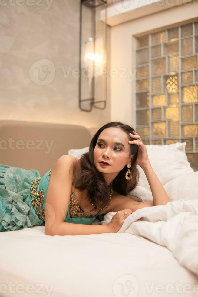 een mooi Aziatisch vrouw is ontspannende Aan een wit bed terwijl vervelend bedenken en jurk foto