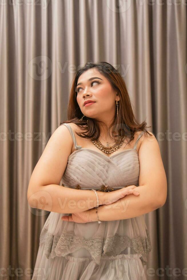 een Aziatisch vrouw vervelend een grijs jurk en mooi bedenken terwijl staand in voorkant van luxueus gordijnen Bij een partij foto