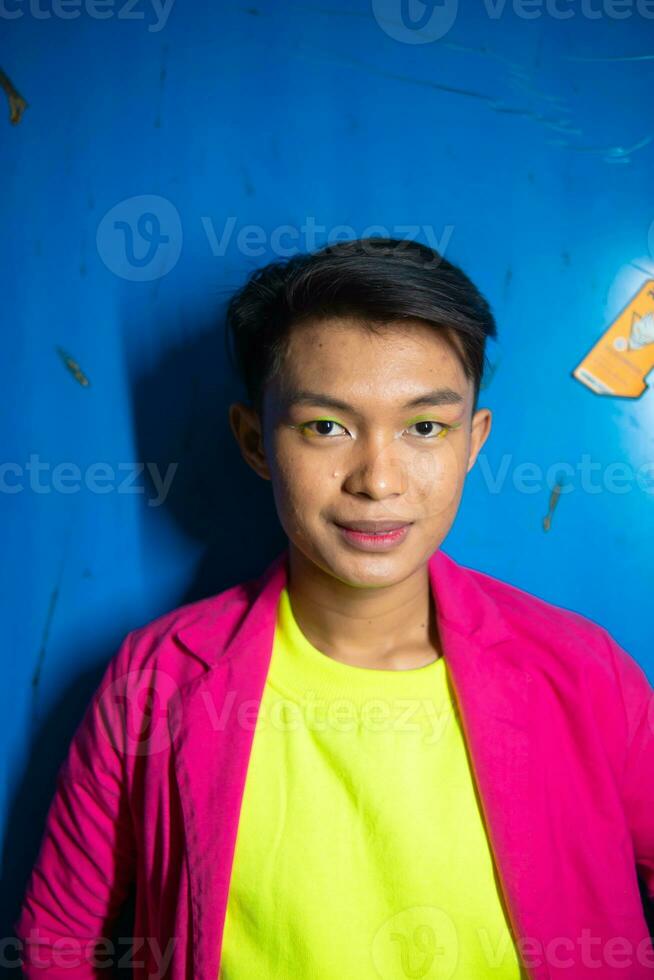 een homo Aziatisch Mens poseren heel grappig en humoristisch terwijl vervelend een roze pak in voorkant van een blauw achtergrond foto