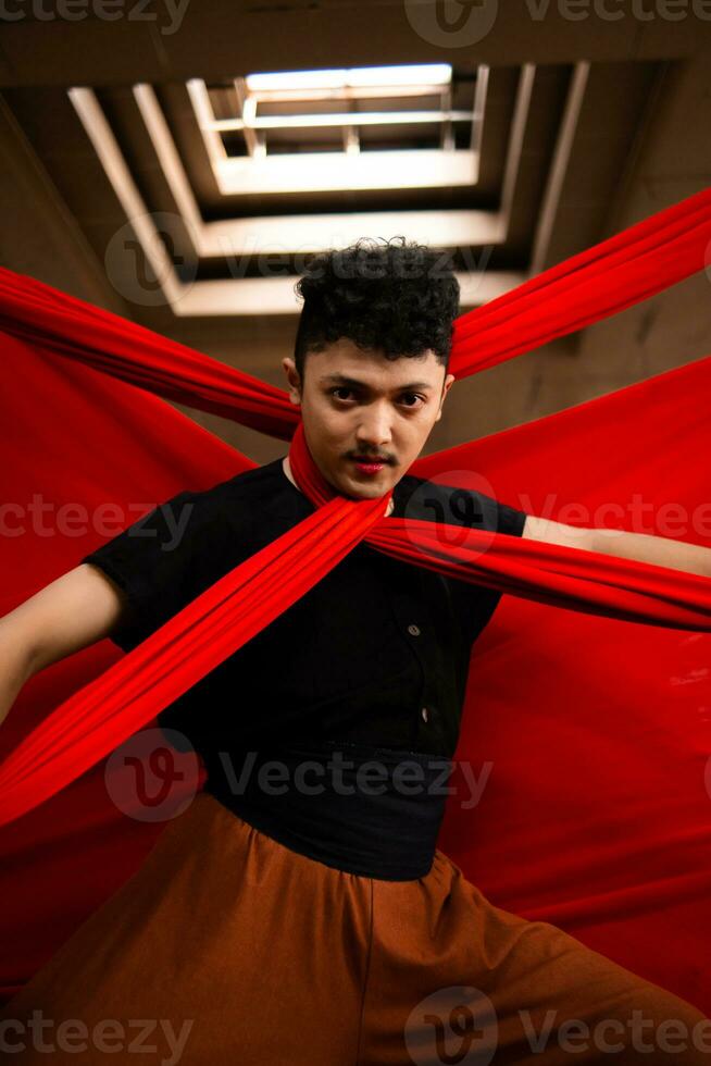 een Aziatisch Mens in een zwart overhemd is gewurgd met een rood kleding terwijl staand in een oud gebouw tegen een rood achtergrond foto