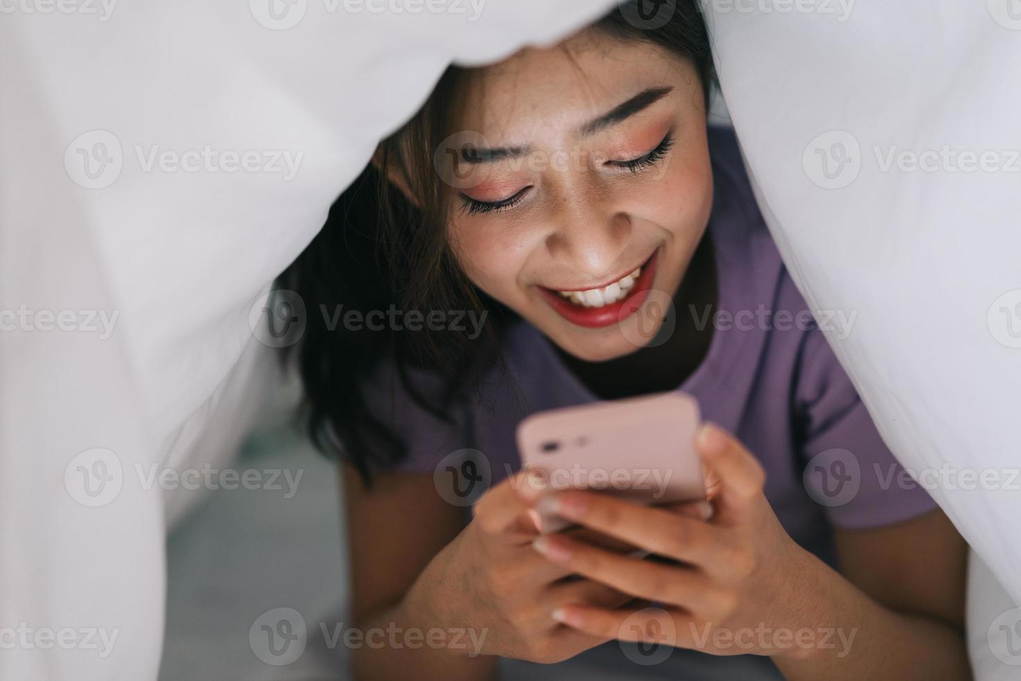 jong Aziatisch meisje dat in deken ligt en haar telefoon gebruikt om te sms'en met vrienden op sociale netwerken foto