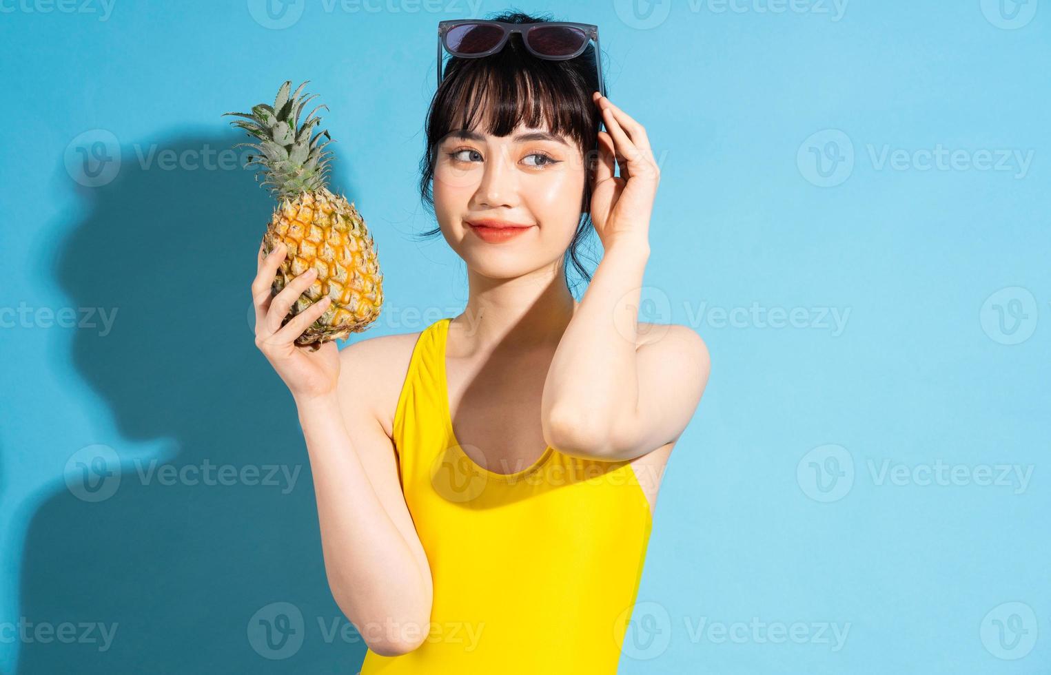 mooie aziatische vrouw die gele jumpsuit op blauwe achtergrond draagt en tropisch fruit eet, zomerconcept foto