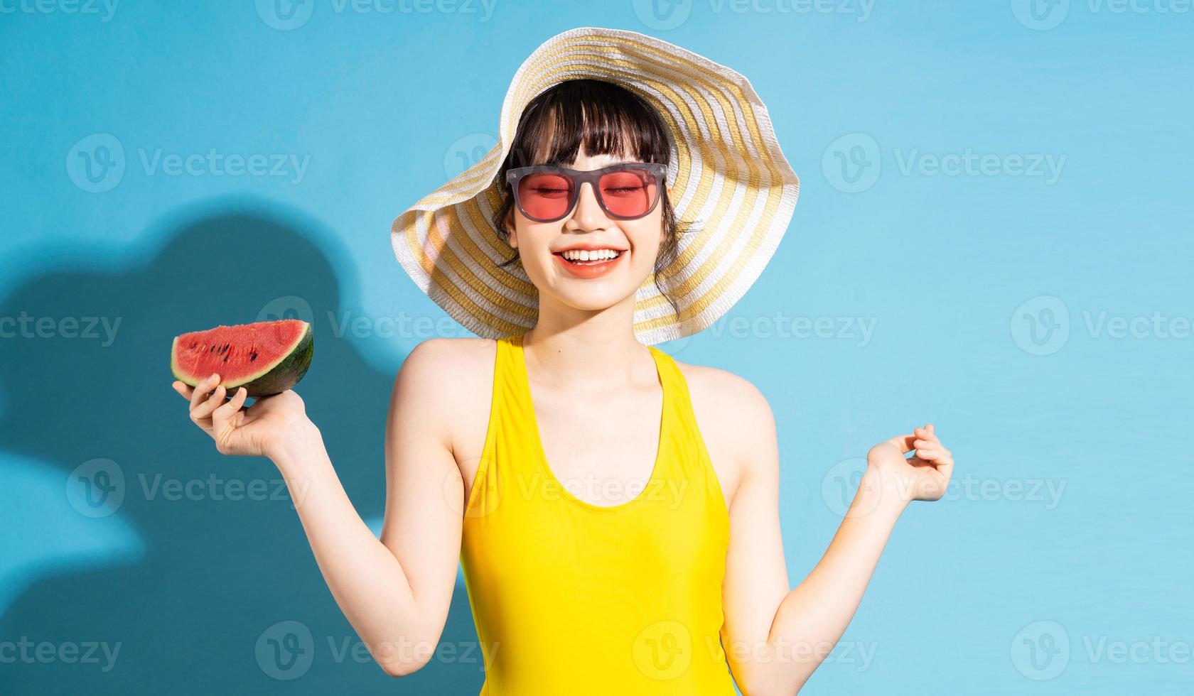 mooie aziatische vrouw die gele jumpsuit op blauwe achtergrond draagt en tropisch fruit eet, zomerconcept foto