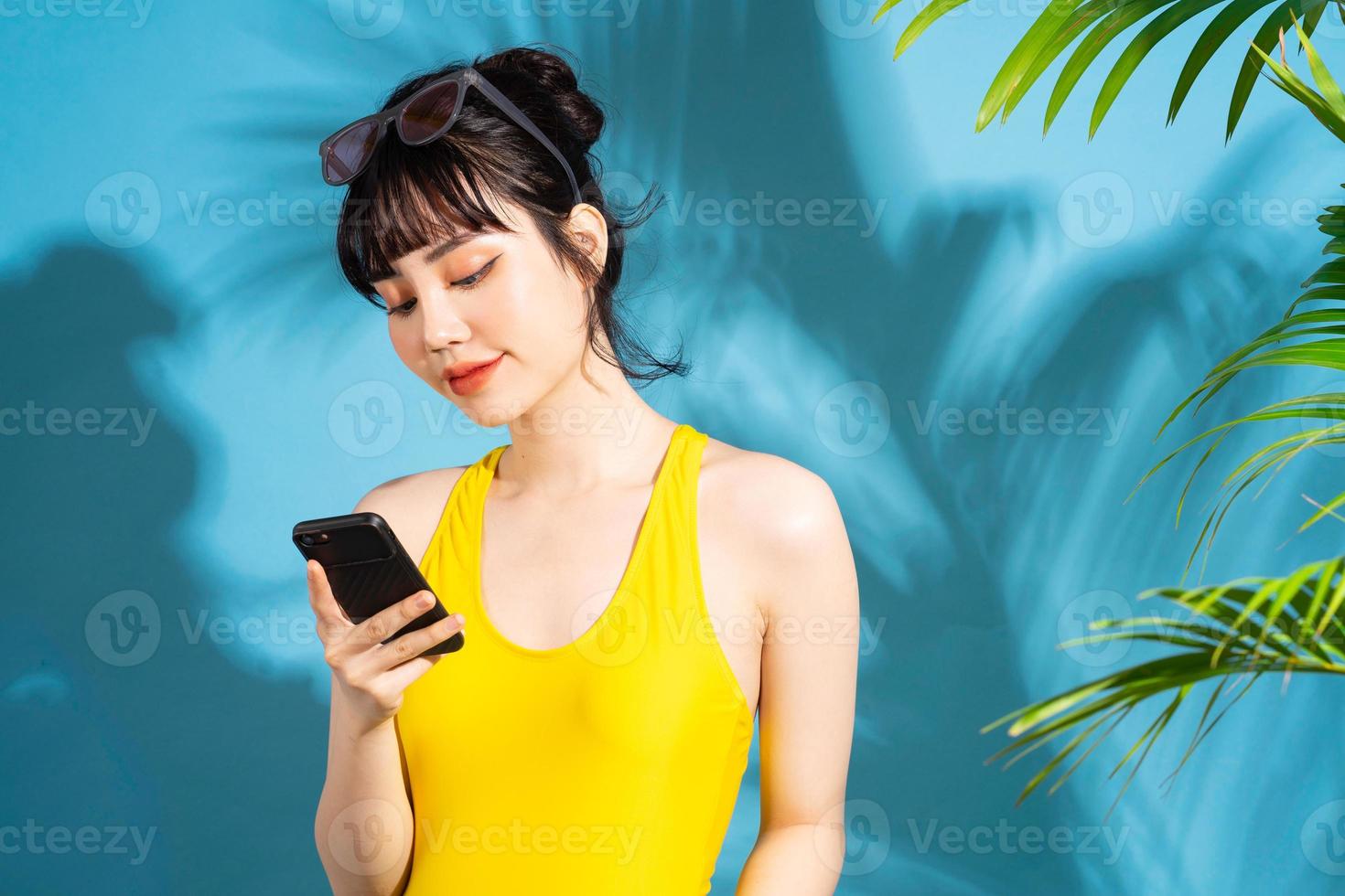 mooie aziatische vrouw die gele jumpsuit op blauwe achtergrond draagt en smartphone gebruikt, zomerconcept foto