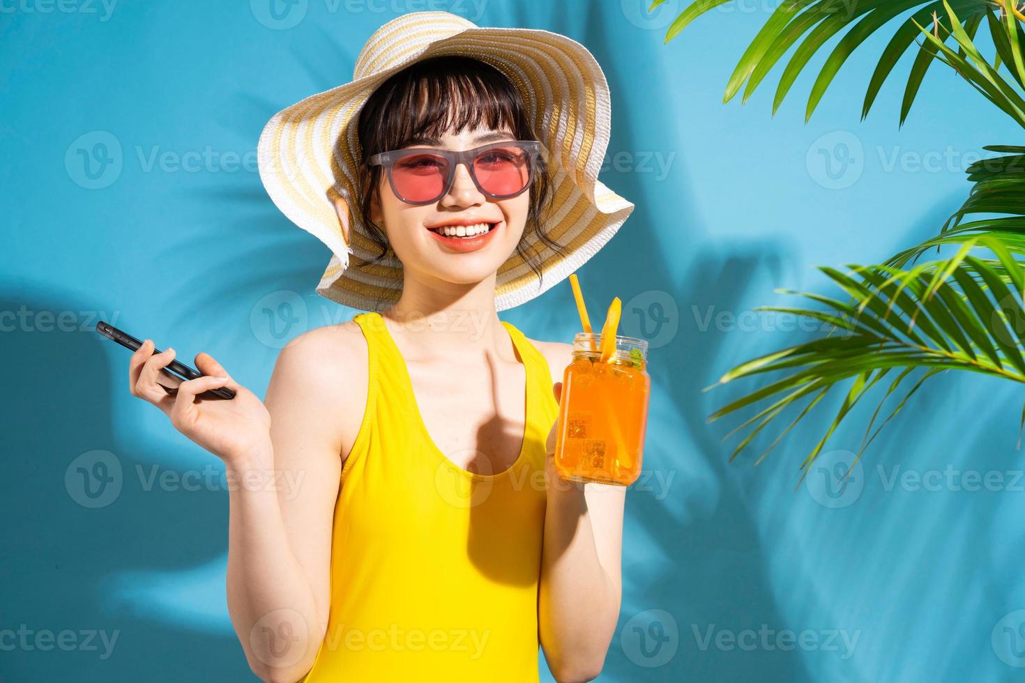 mooie aziatische vrouw die gele jumpsuit op blauwe achtergrond draagt en sap drinkt, zomerconcept foto