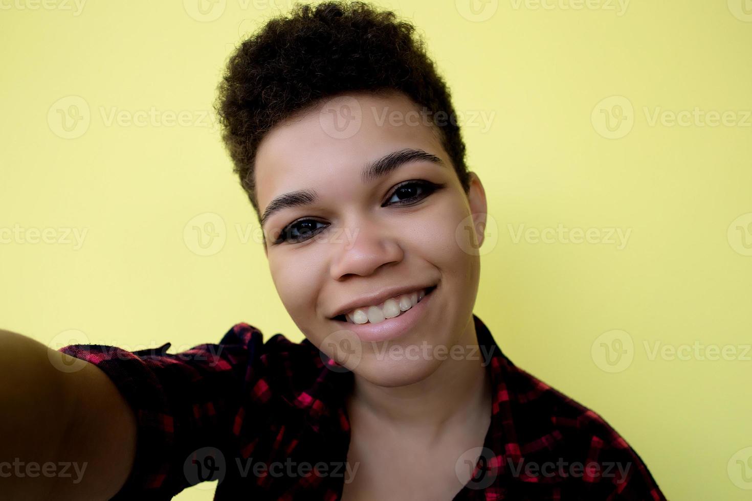 mooie en gelukkige Afro-Amerikaanse vrouw met kort haar op een gele achtergrond, neemt een selfie, close-up portret foto