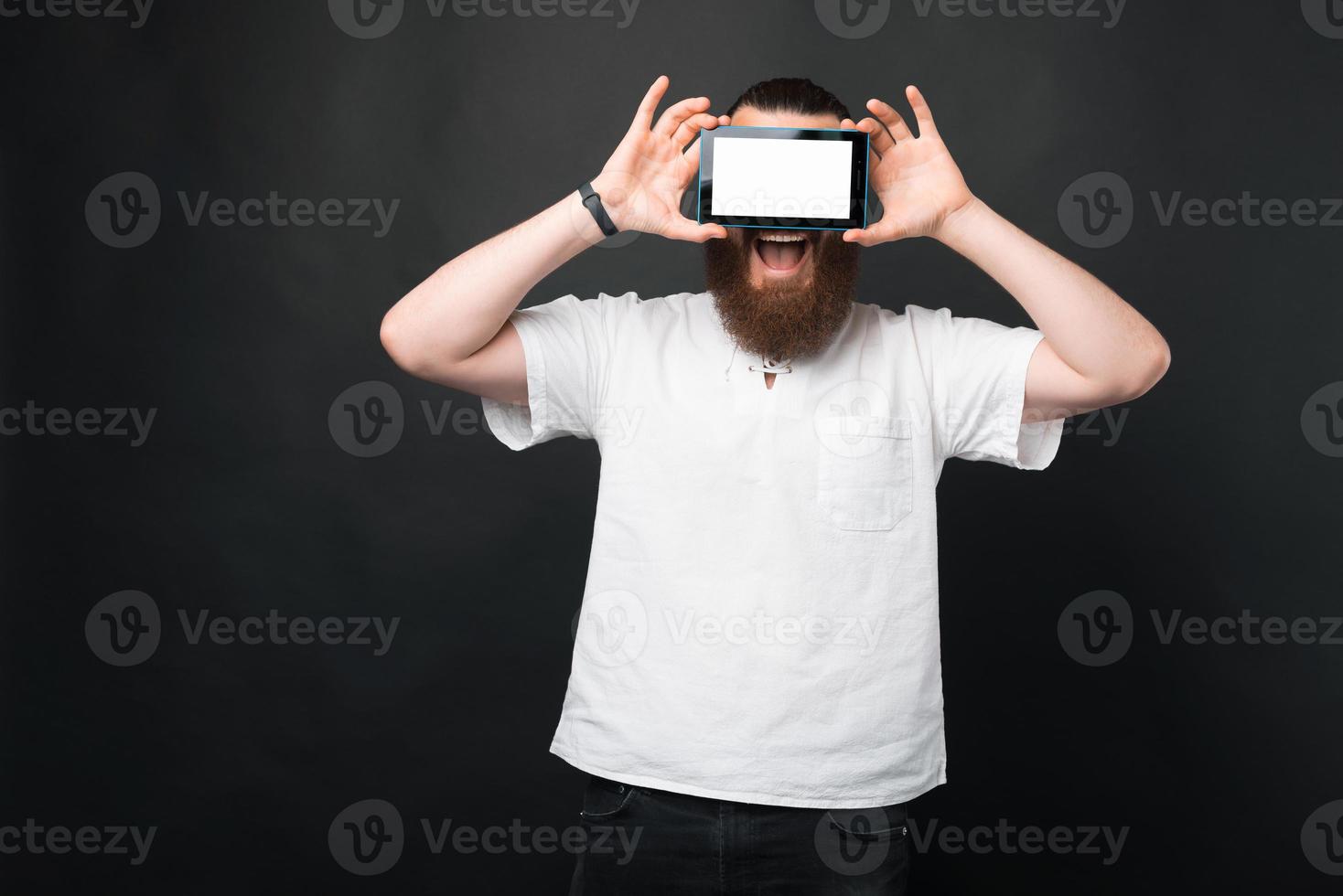 portret van een verbaasde bebaarde man die een tablet boven de ogen houdt met een wit scherm foto