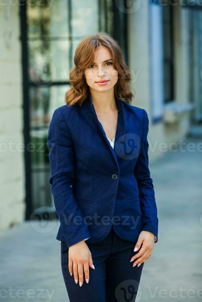 portret van een mooi jong vrouw met lang bruin haar, gekleed in een blauw jasje, staand tegen de achtergrond van een kantoor gebouw. portret van jong bedrijf vrouw gebruik makend van mobiel telefoon foto