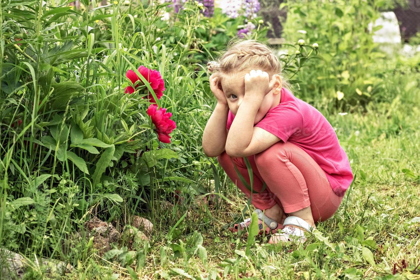 een klein meisje met haar hoofd in gedachten, zittend bij een bloembed in de tuin met roze pioenrozen foto