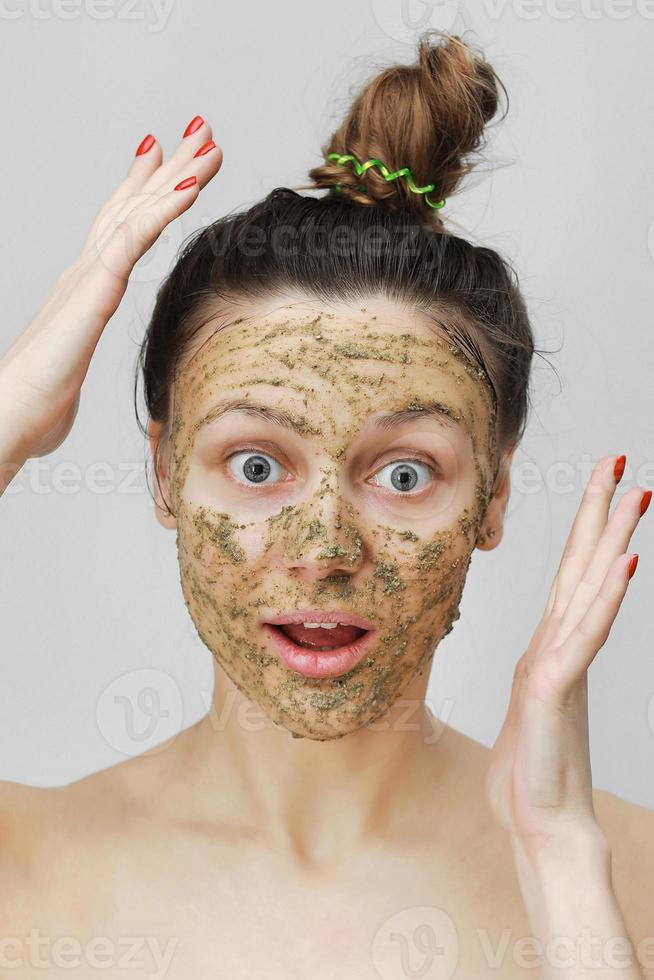 huidverzorging. cosmetische dag. verbaasd jong meisje in huisstijl, haar haar verzameld met haar handen bovenaan. met eco, kruiden, natuurlijk masker, groen op het gezicht. vooraanzicht foto