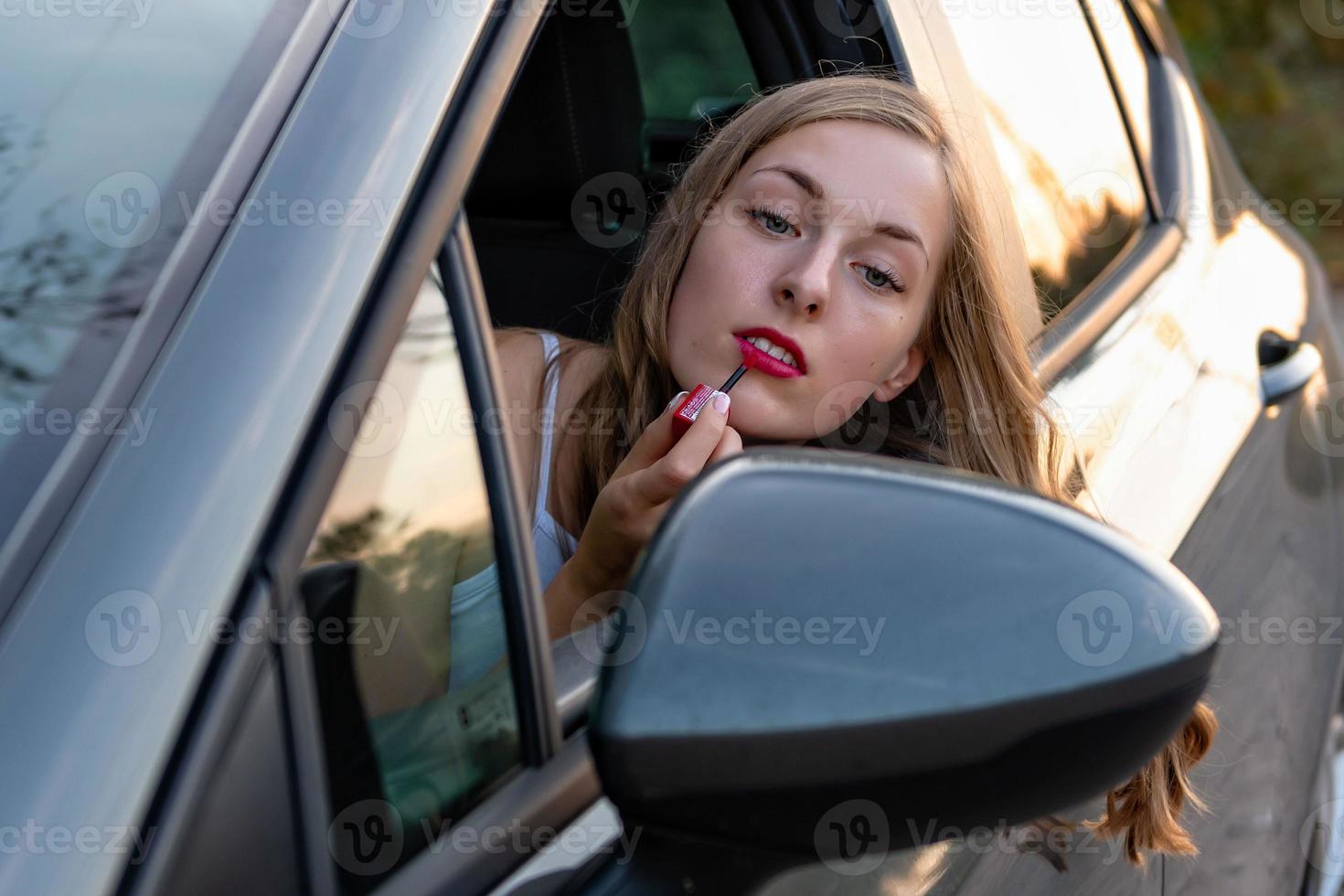 een jonge, mooie vrouw met lang haar door het autoraam kijkt in de achteruitkijkspiegel en schildert haar lippen. foto