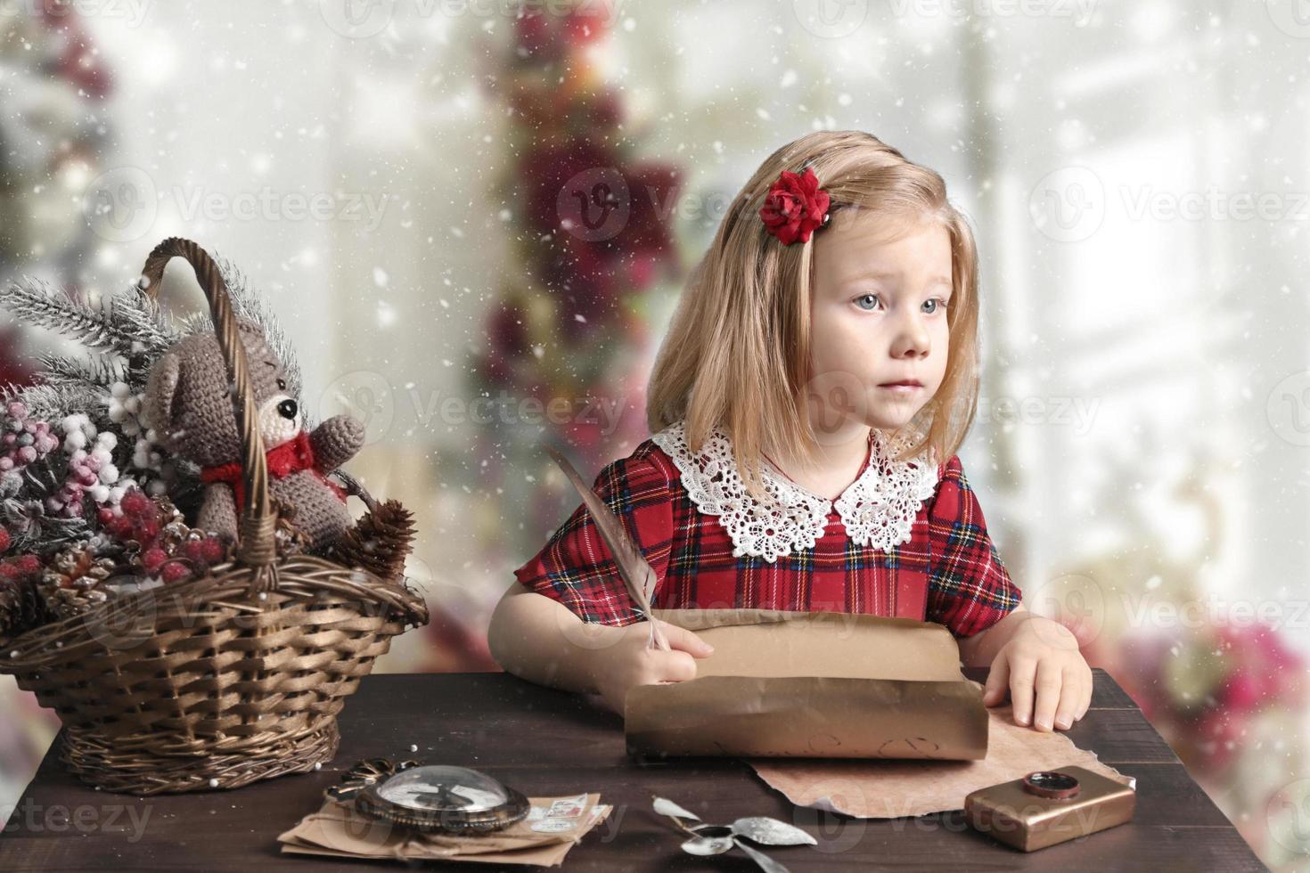 een klein meisje in een rode jurk zit aan de tafel en schrijft een brief aan de kerstman. kerstkaart foto