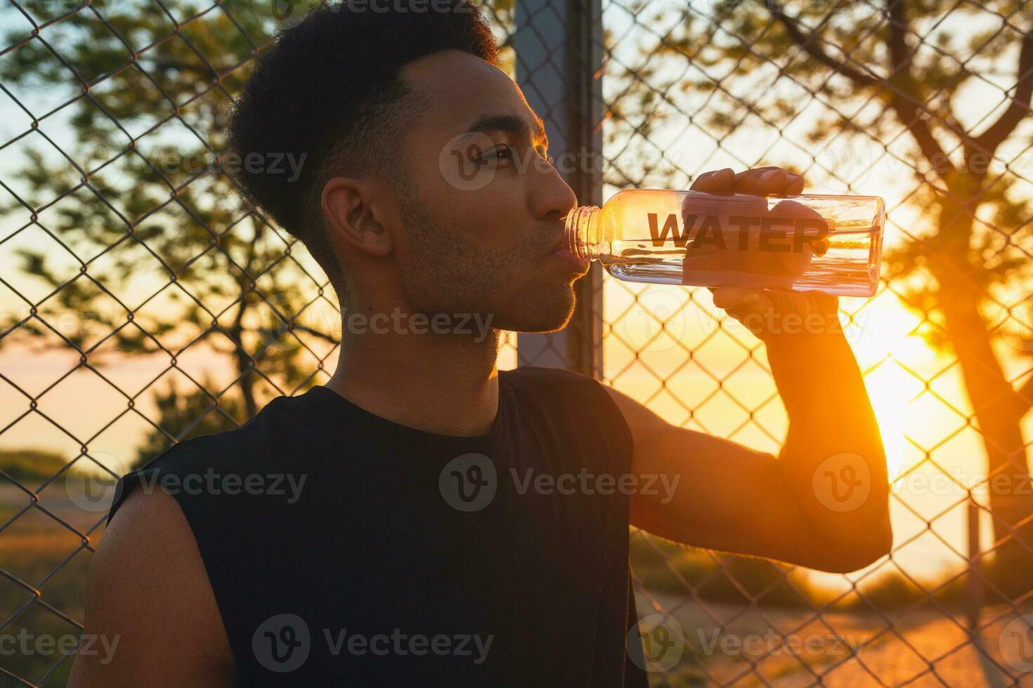 zwart Mens aan het doen sport- in ochtend, drinken water Aan basketbal rechtbank Aan zonsopkomst foto