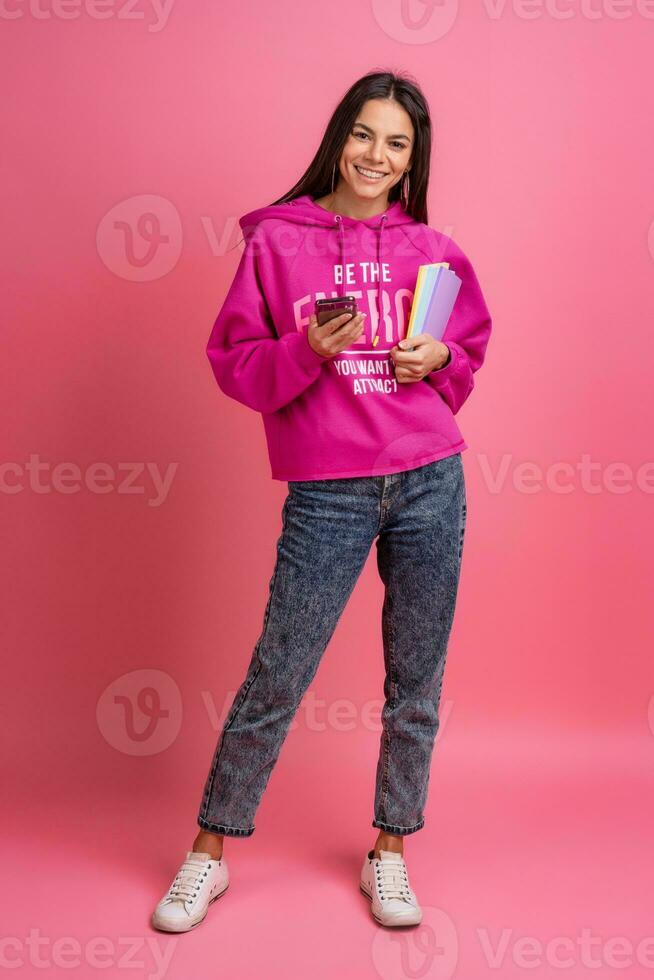 spaans mooi vrouw in roze capuchon glimlachen Holding Holding notebooks en gebruik makend van smartphone foto