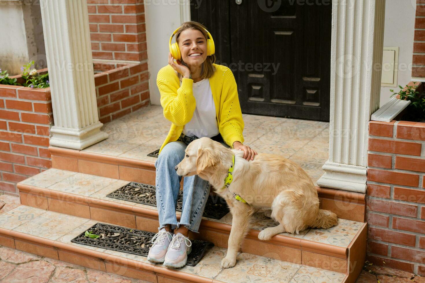 vrouw in geel trui wandelen Bij haar huis met een hond luisteren naar muziek- in hoofdtelefoons foto