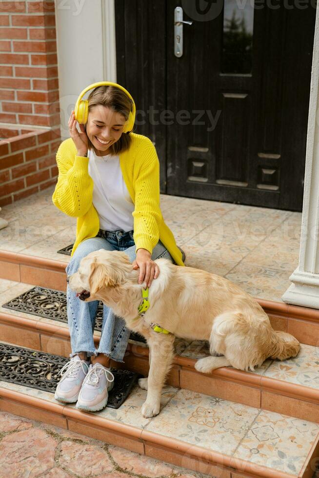 vrouw in geel trui wandelen Bij haar huis met een hond luisteren naar muziek- in hoofdtelefoons foto