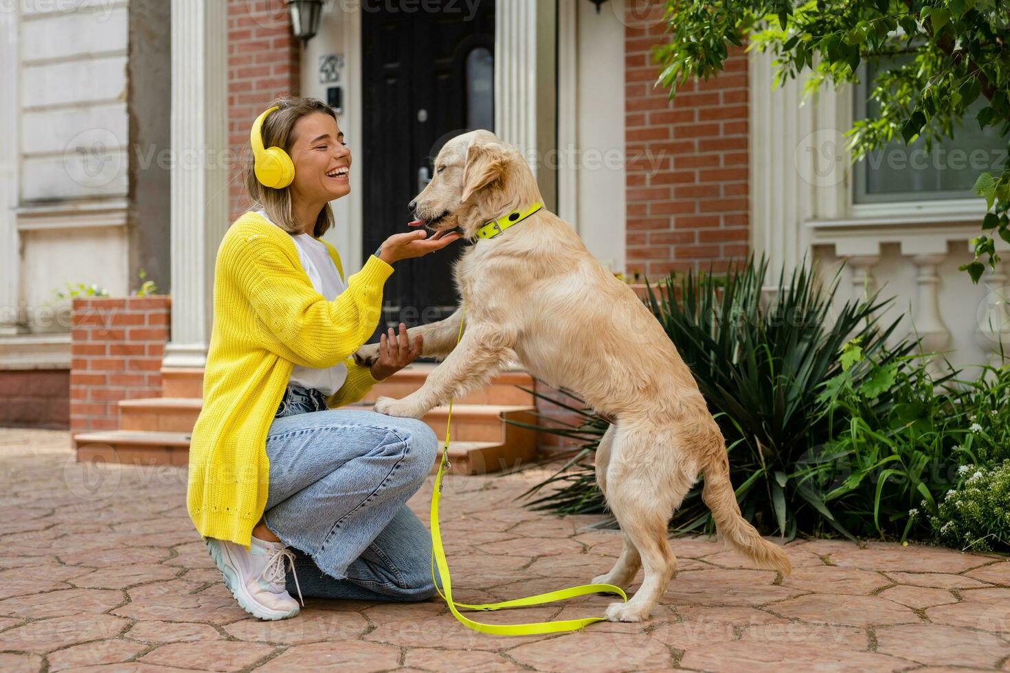 vrouw in geel trui wandelen Bij haar huis met een hond luisteren naar muziek- in hoofdtelefoons foto