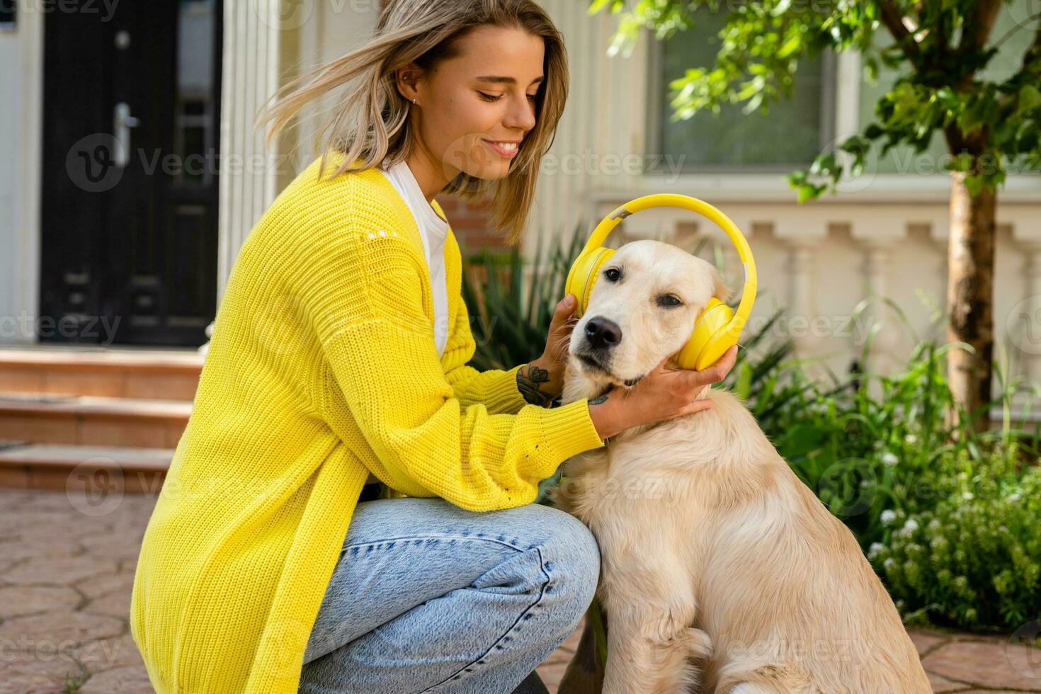 vrouw in geel trui wandelen Bij haar huis met een hond luisteren naar muziek- in hoofdtelefoons foto