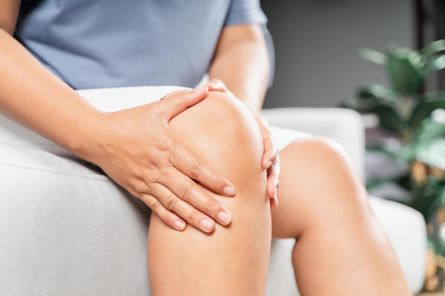 close-up van een mollige vrouw die op de bank zit en kniepijn voelt en ze masseert haar knie. gezondheidszorg en medisch concept. foto