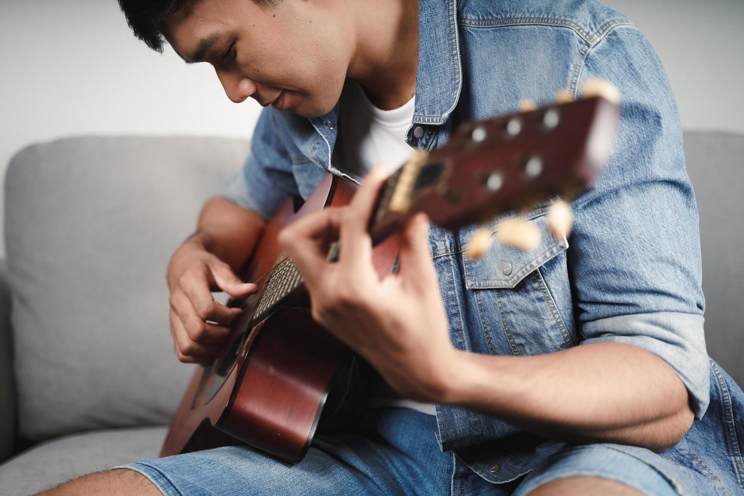 geniet van knappe aziatische man die gitaar oefent of speelt op de bank in de woonkamer living foto