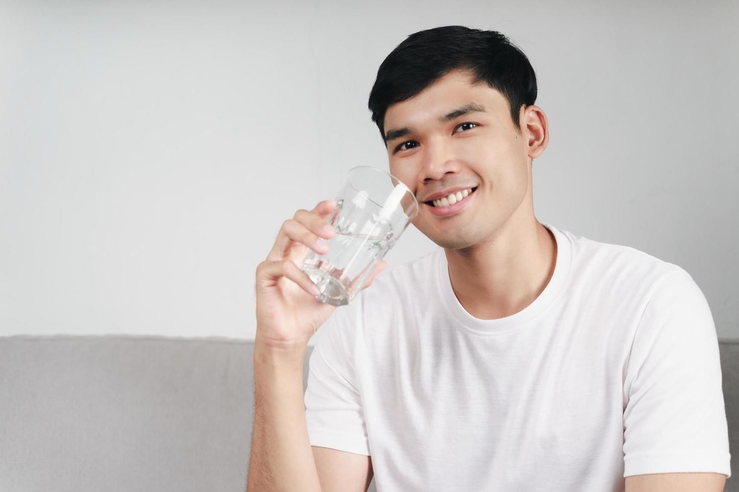 knappe aziatische man die een glas water drinkt op de bank in de woonkamer foto