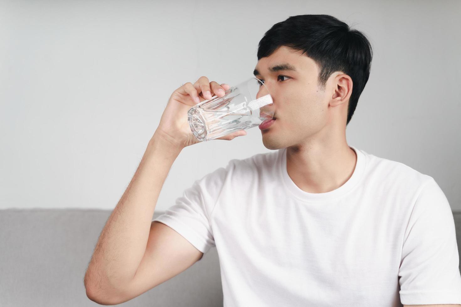 knappe aziatische man die een glas water drinkt op de bank in de woonkamer foto