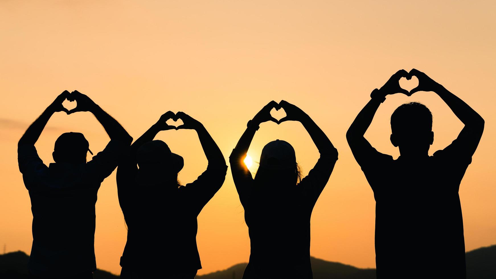 groep mensen met opgeheven armen en hand naar de hartvorm kijkend naar zonsopgang op de bergachtergrond. geluk, succes, vriendschap en communautaire concepten. foto