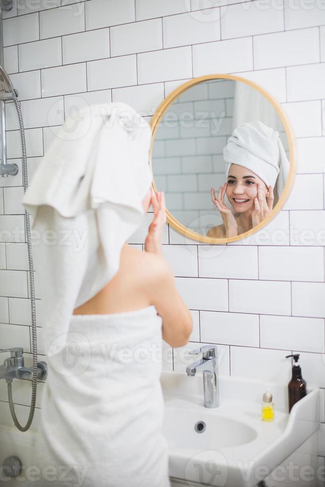 jonge vrouw in witte handdoek brengt vochtinbrengende olie aan op de gezichtshuid. foto