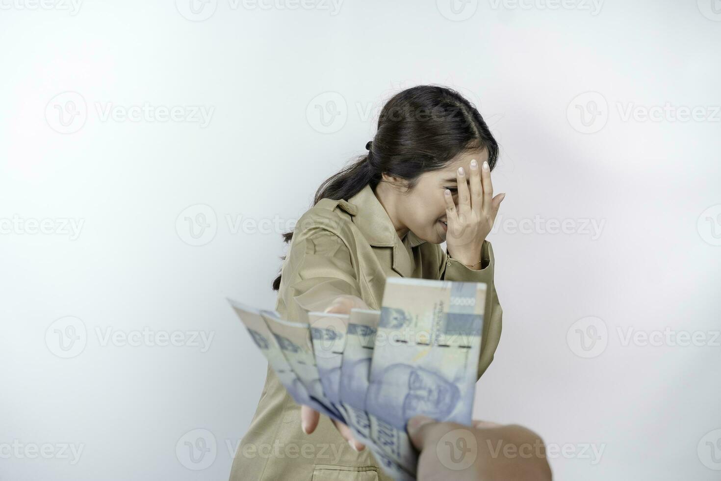 een echt jong regering arbeider vrouw is vervelend khaki uniform gebaren afwijzing naar contant geld geld in Indonesisch roepia geïsoleerd door wit achtergrond foto