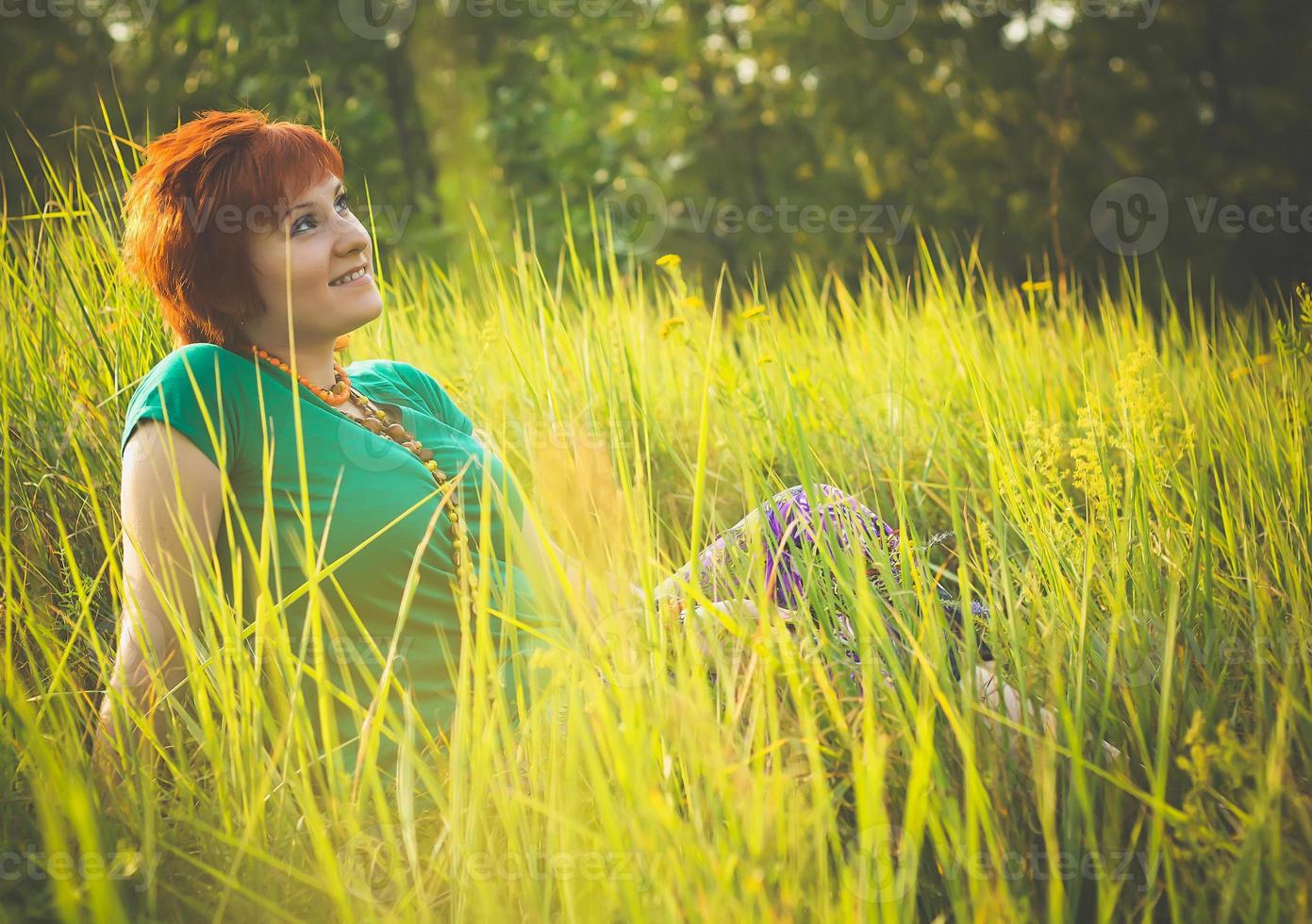 jonge roodharige vrouw zittend in het gras en genieten van zonnige zomerdag. portret van gelukkig lachend jong meisje in de natuur. foto