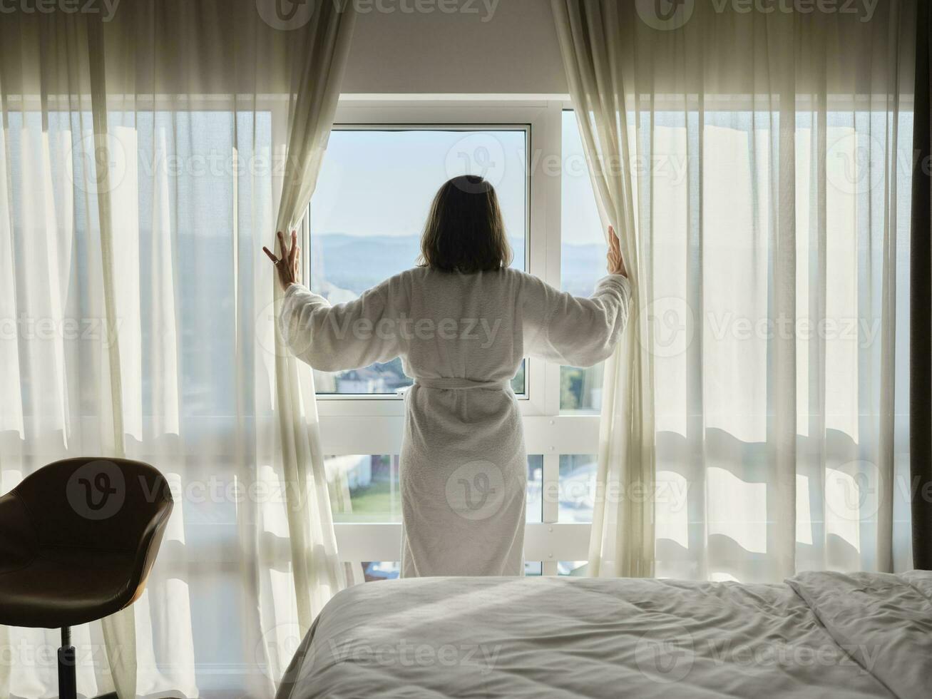 vrouw opening wit gordijn terwijl op zoek door venster Bij luxe hotel kamer foto