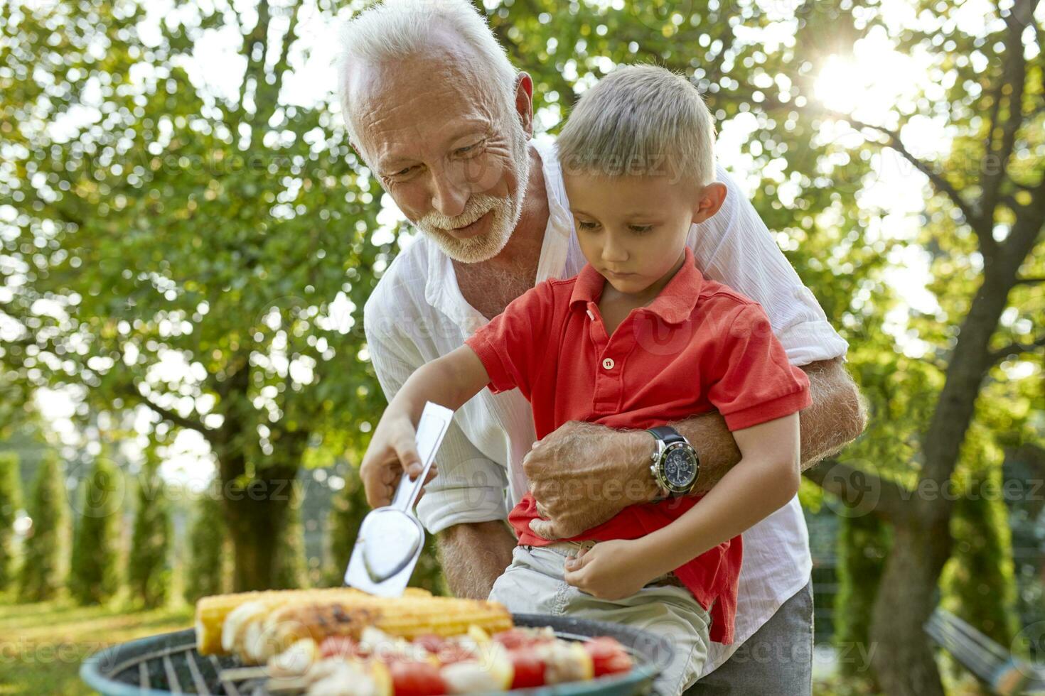 opa helpen kleinzoon draaien een maïs maïskolf gedurende een barbecue in tuin foto