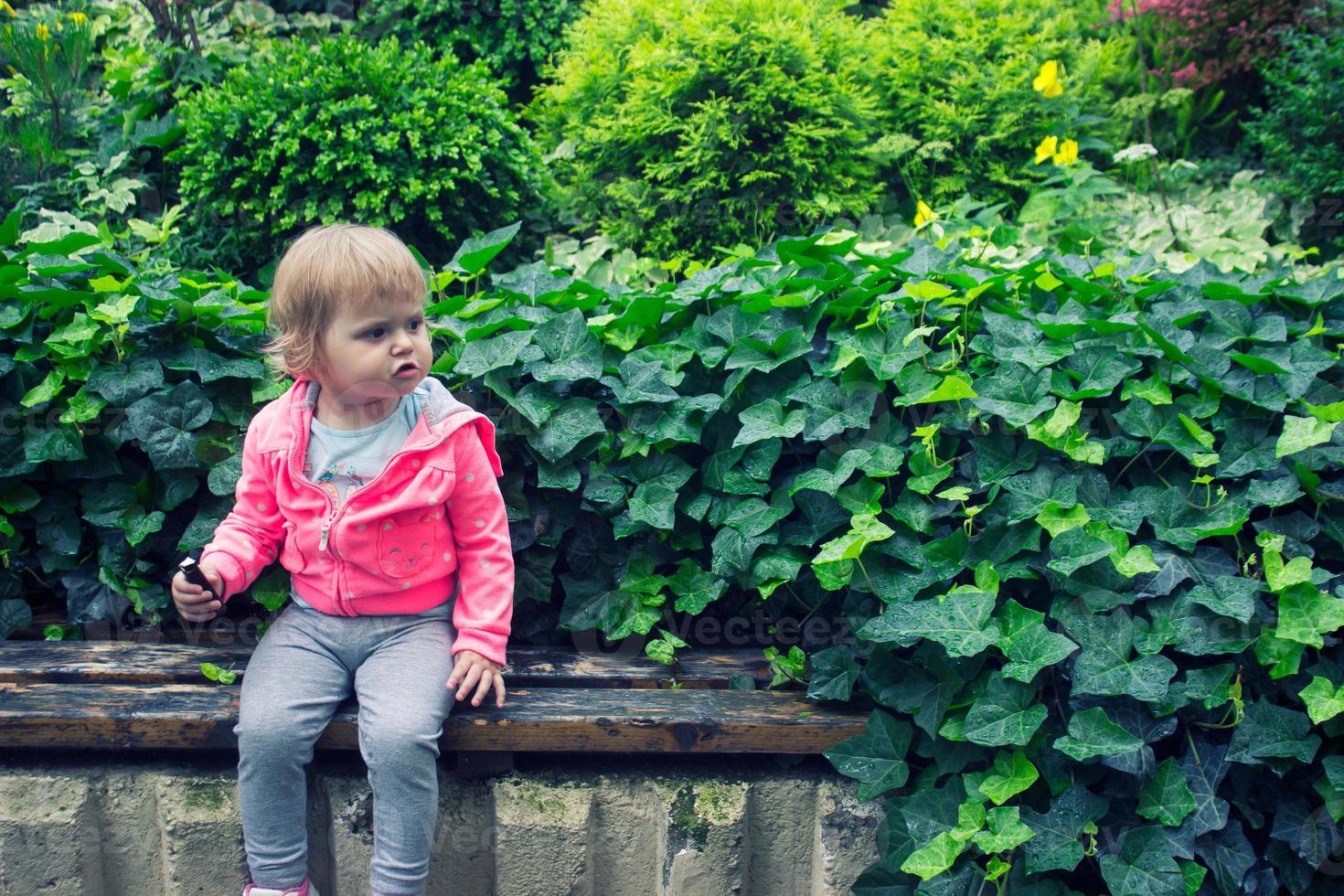 klein meisje zit in de tuin, zittend op de bank snoep te eten foto